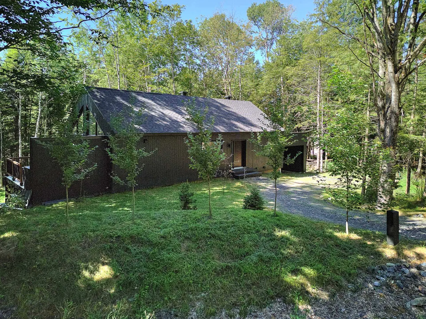 A modern brown house with a gray roof is surrounded by green trees and grass on a sunny day.