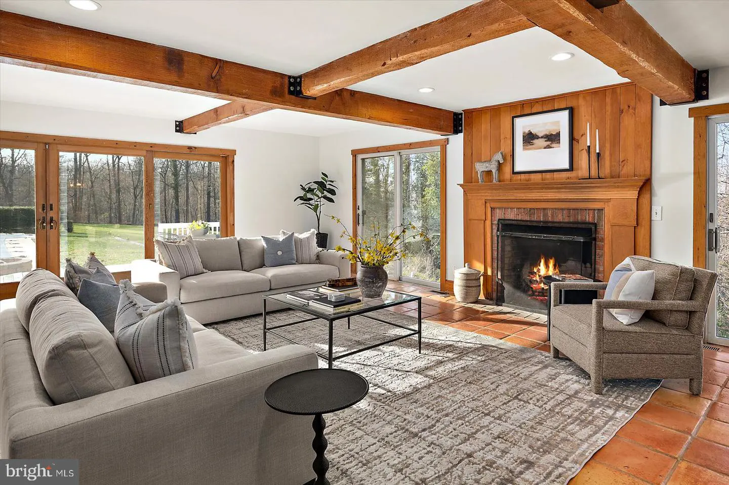 Living room with exposed wood beams, sofas, armchair, and fireplace. Large windows overlook a green lawn.