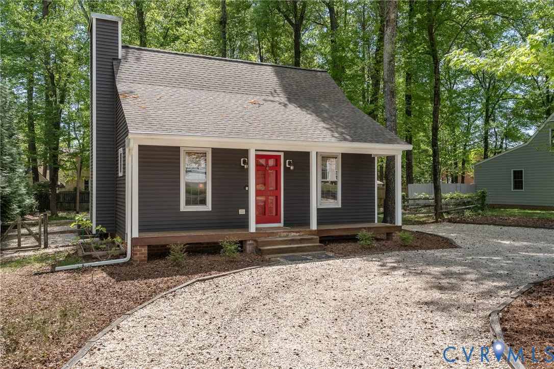 A gray house with a red door, white trim, and a gravel driveway is surrounded by trees.