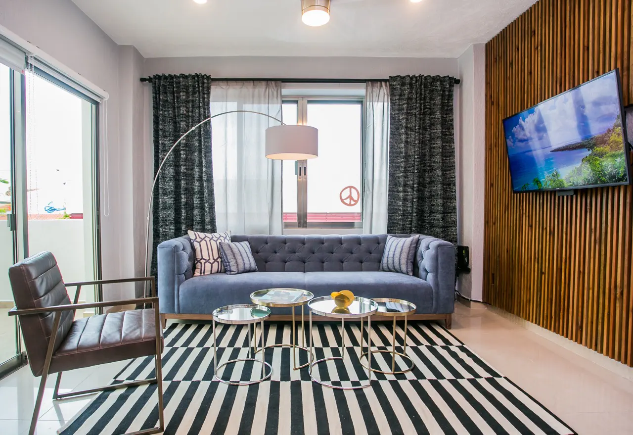 Living room with blue sofa, striped rug, and wood-paneled wall with a TV. A modern chair sits near a sliding glass door.