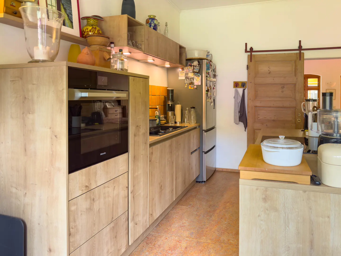A bright kitchen with light wood cabinets, stainless steel fridge, and a sliding barn door.