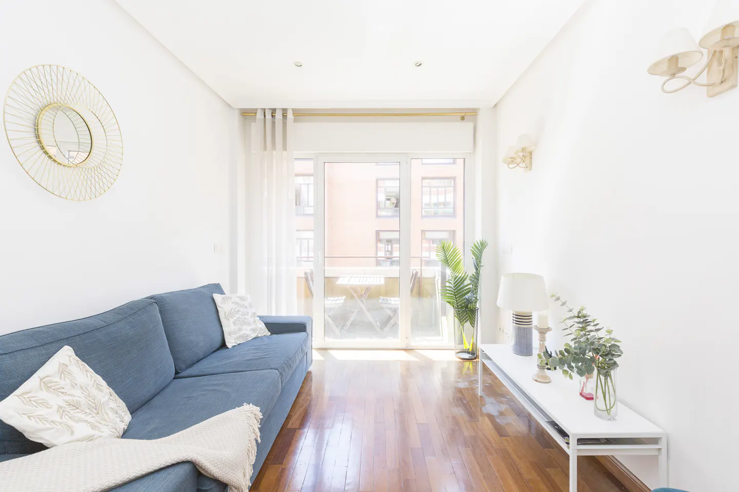 Bright living room with a blue sofa, white walls, and hardwood floors. A balcony is visible through glass doors.