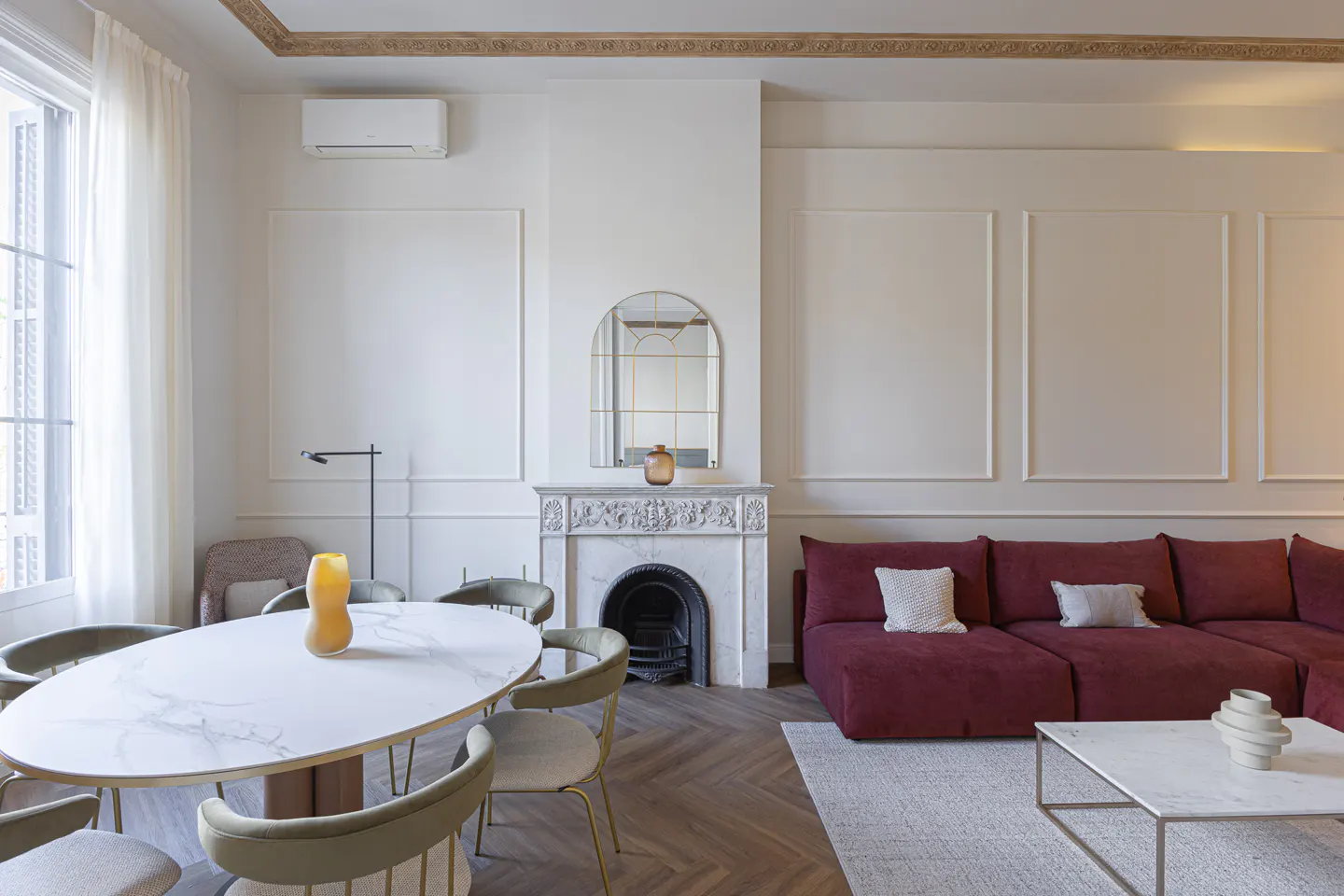 Bright living room with white walls, herringbone floors, and a burgundy sectional sofa. A marble fireplace and oval dining table add elegance.