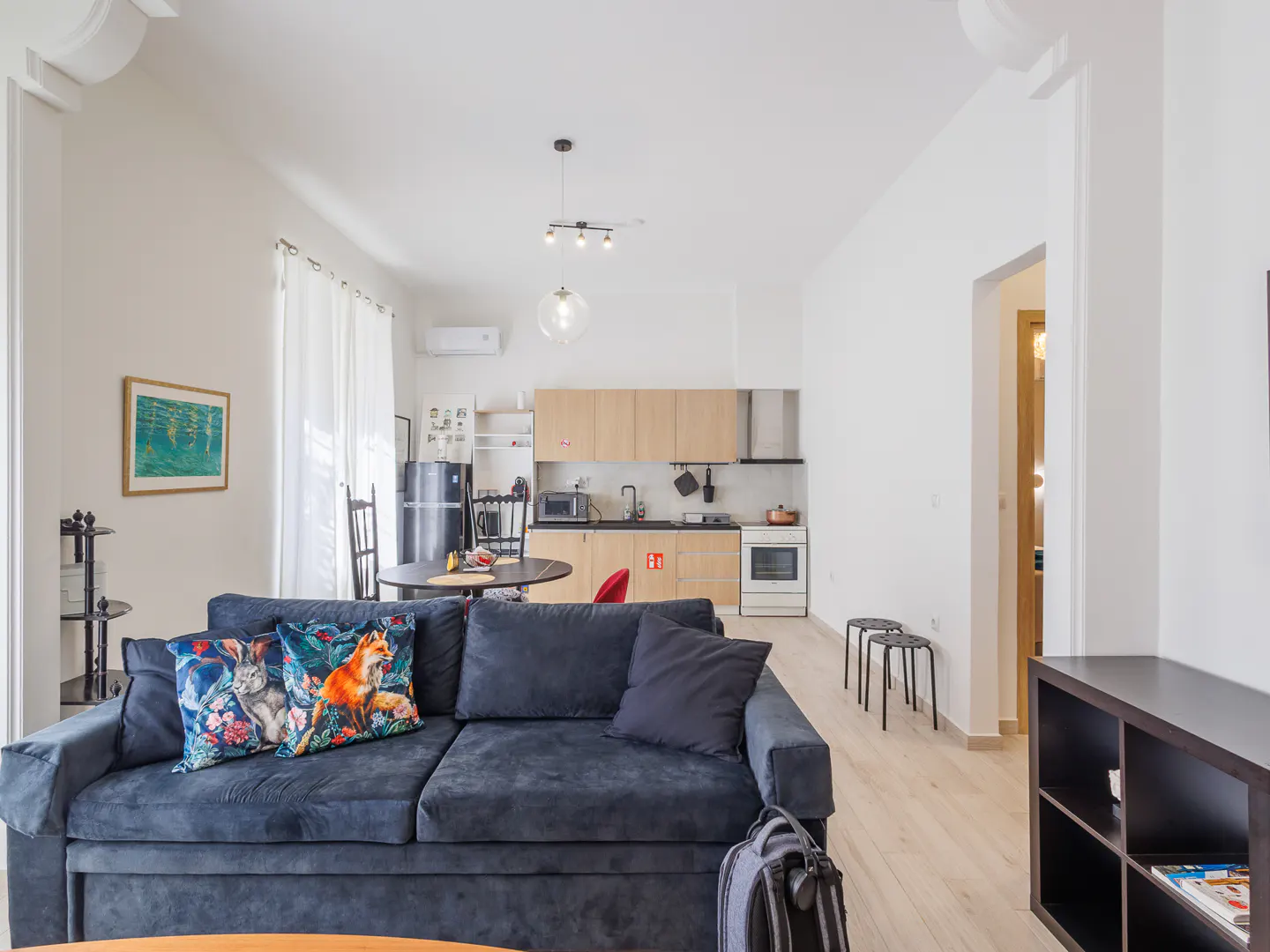 Bright apartment interior with a blue sofa, animal print pillows, and a kitchen in the background. Light wood floors and white walls.