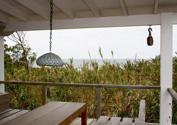 Covered wooden deck with a dining table, hanging lamp, and view of tall coastal grasses and the ocean in the distance.