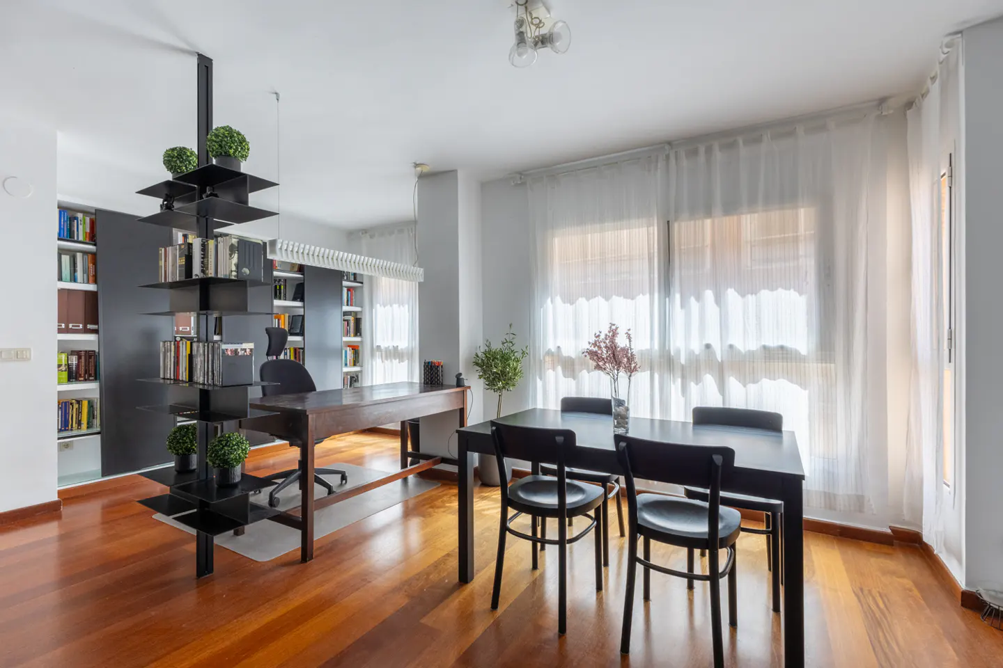 Bright room with wood floors, black table and chairs, and a black bookshelf. White curtains cover the windows.