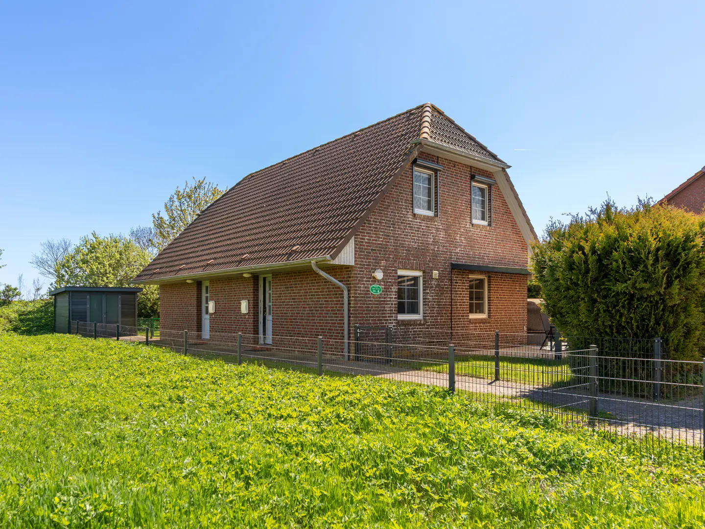 A two-story brick house with a brown roof sits on a green lawn under a blue sky. A small shed is to the left of the house.