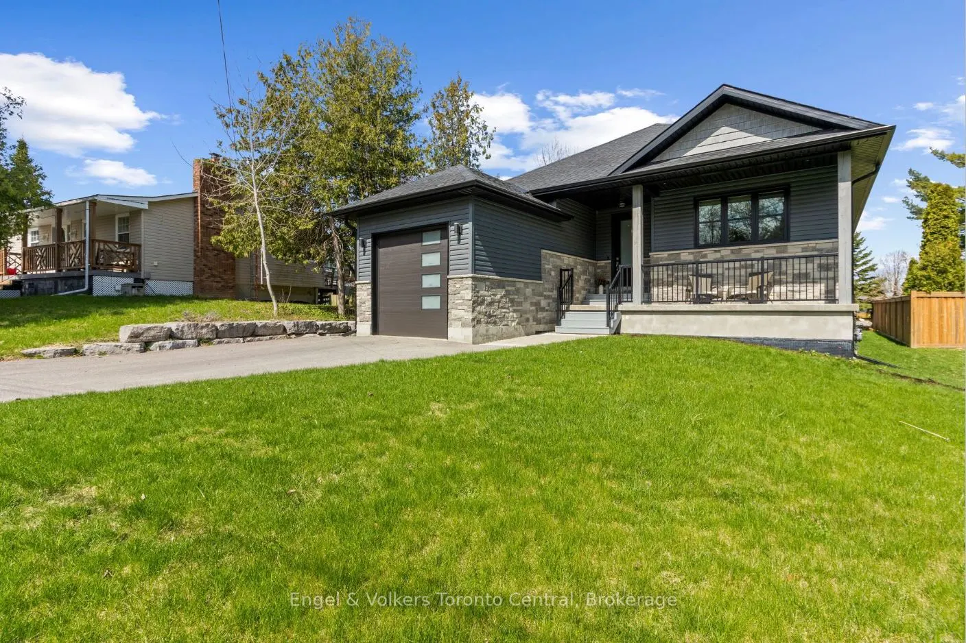 A modern, gray house with a stone foundation and a brown garage door sits on a green lawn under a blue sky.
