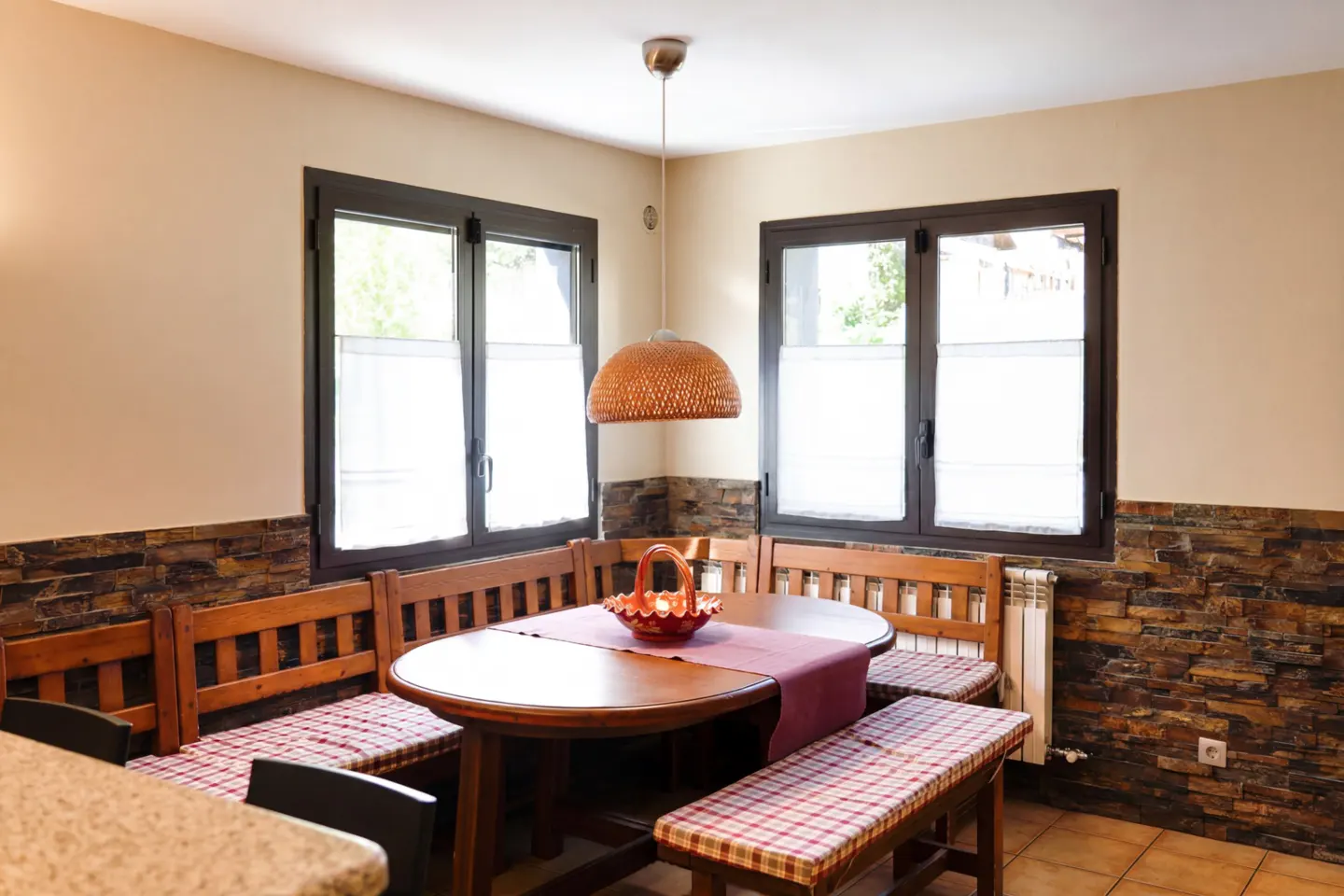 Cozy dining nook with wood table, benches with red plaid cushions, and woven pendant light. Two windows with white curtains.