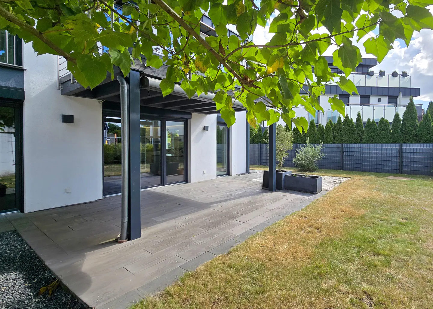 Modern home exterior with a gray patio, white walls, and black trim. Green lawn and trees in the background.