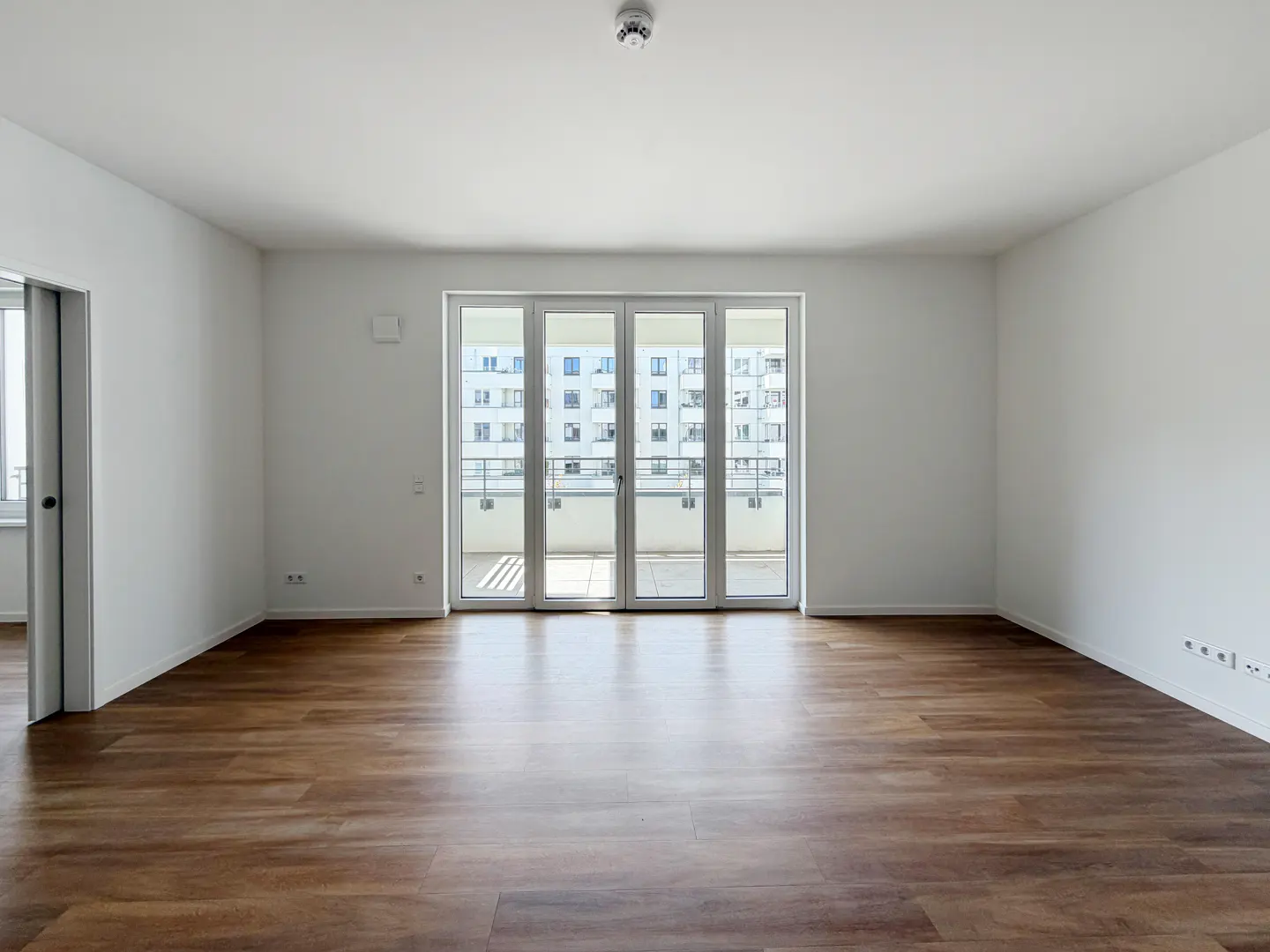 Bright, empty room with wood floors and white walls. Glass doors lead to a balcony with a view of a building.