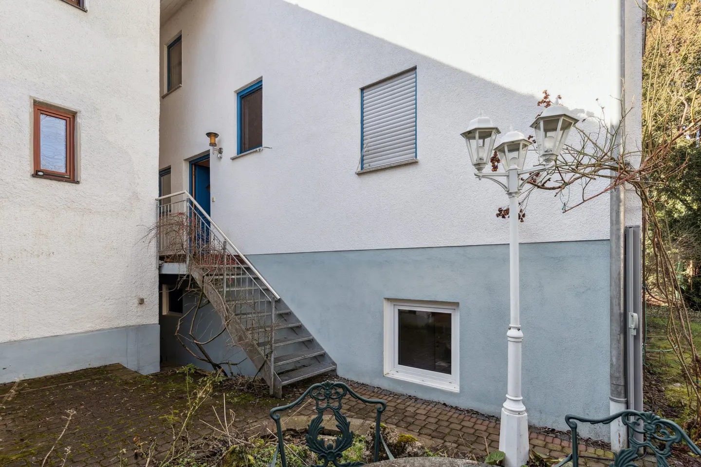 Exterior view of a two-story building with white walls, blue trim, and a metal staircase leading to a blue door. A white lamppost stands nearby.