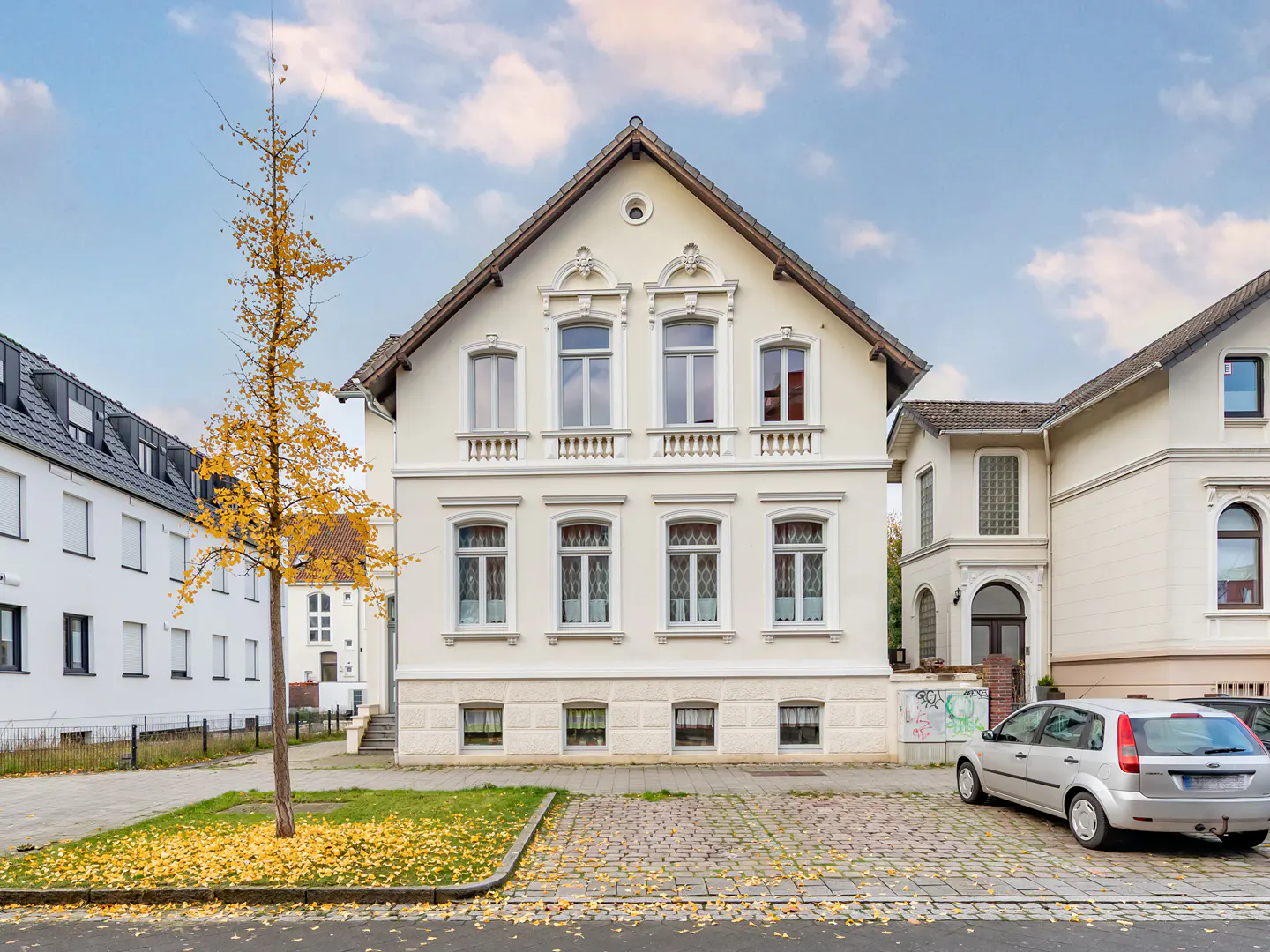 Two-story cream-colored house with a gabled roof, decorative window trim, and a silver car parked in front. A tree with yellow leaves stands to the left.
