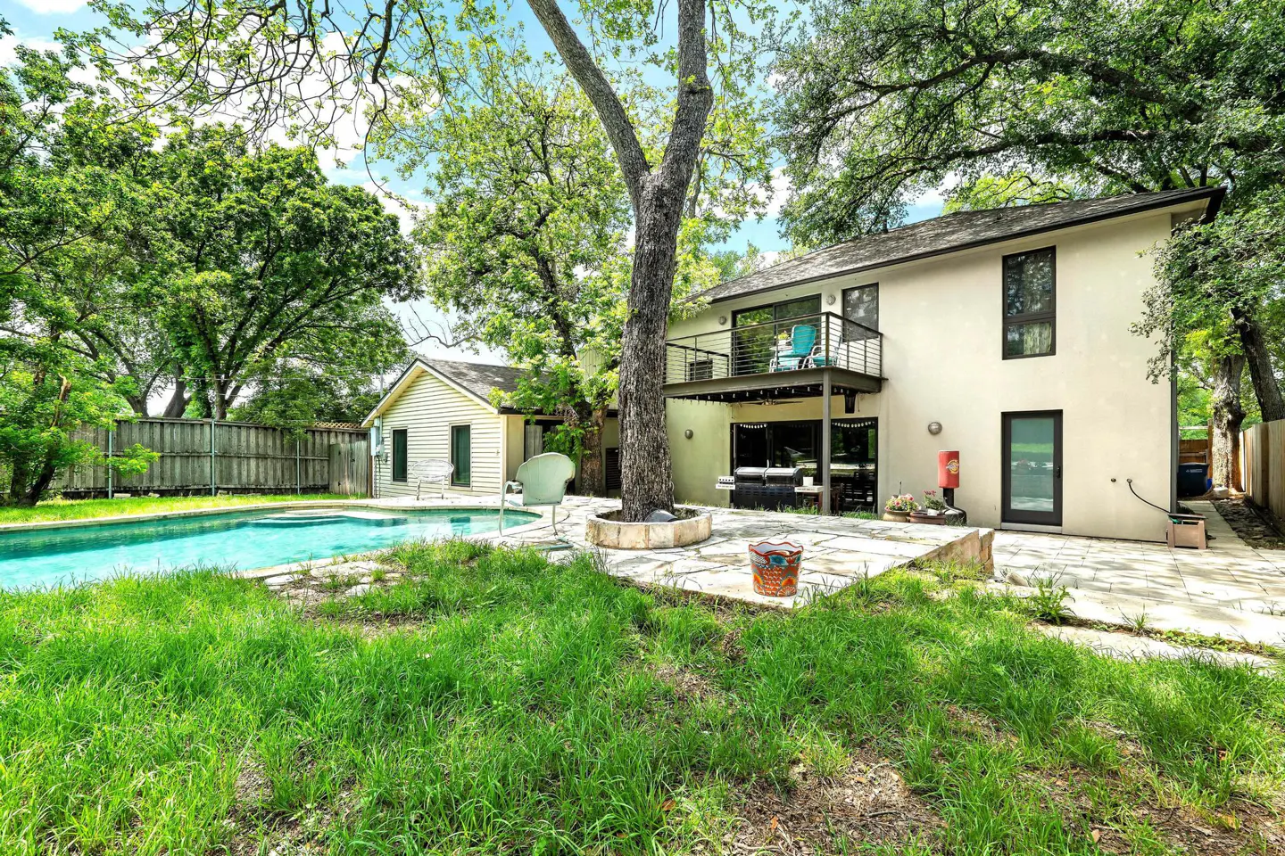 Backyard view of a two-story house with a pool, patio, and lush green lawn. A tree grows through the patio.