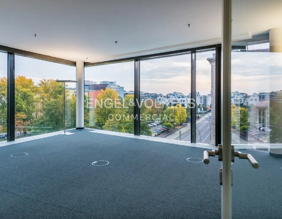 Empty office space with blue carpet and floor outlets. Large windows offer a city view with trees and buildings. Engel & Volkers logo on the glass.