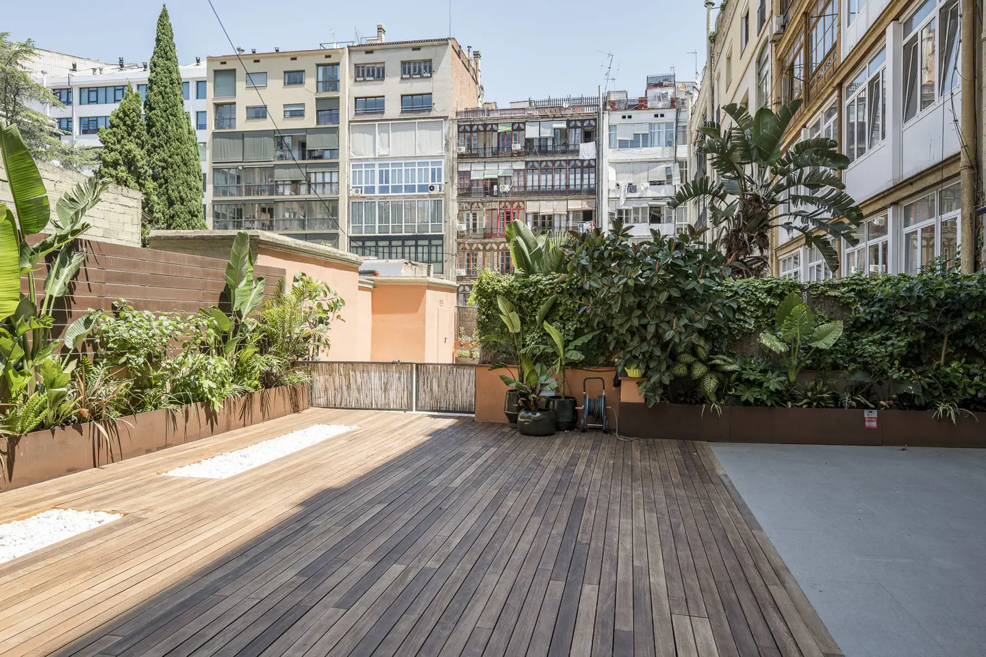 Spacious urban rooftop terrace with wooden decking, lush green plants, and surrounding city apartment buildings.