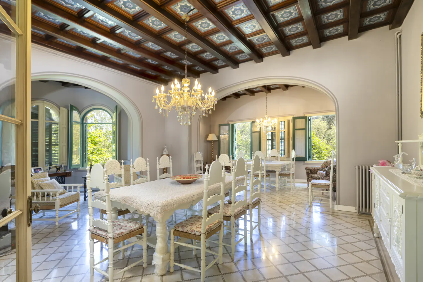 Elegant dining room with a long table, white spindle chairs, crystal chandelier, and ornate coffered ceiling.