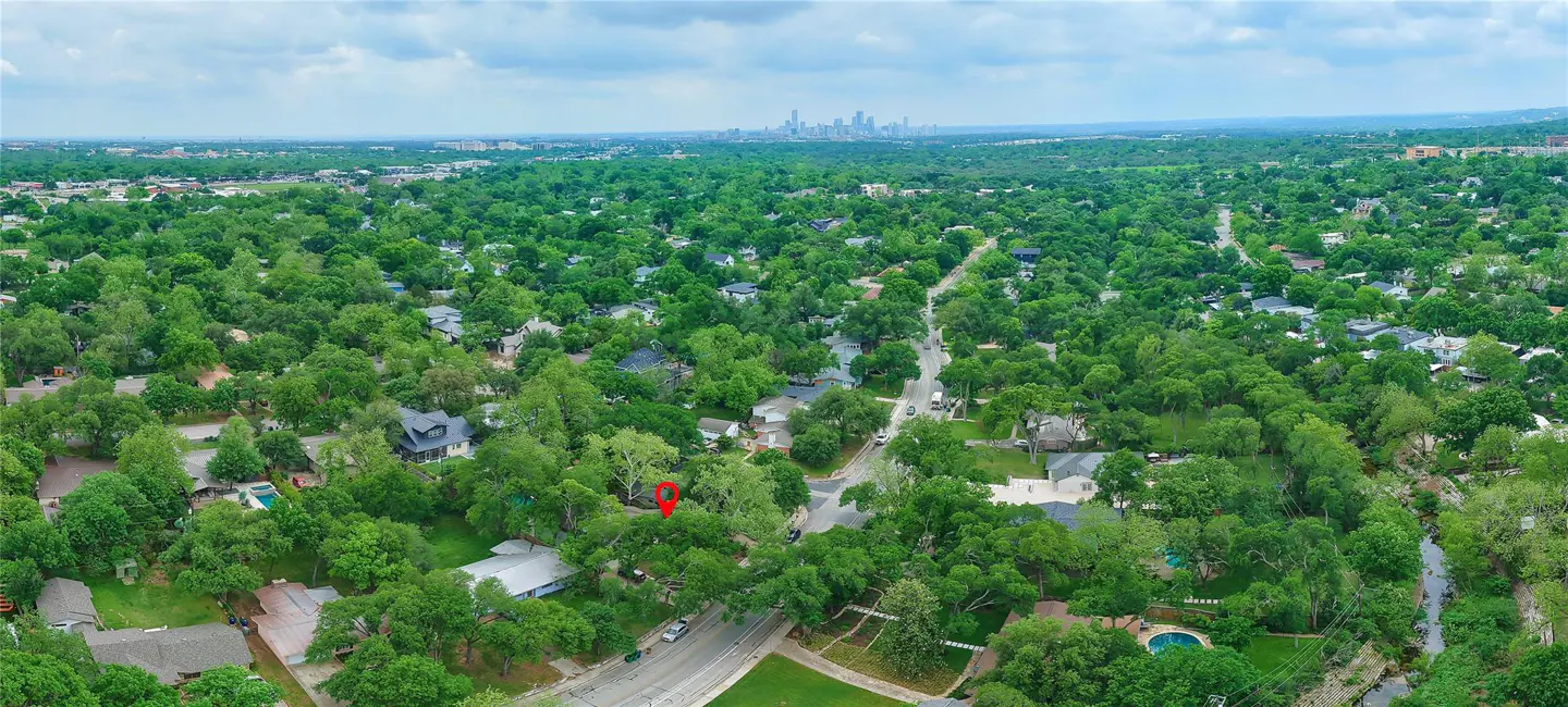 Aerial view of a neighborhood with green trees, houses, roads, and a city skyline in the distance.