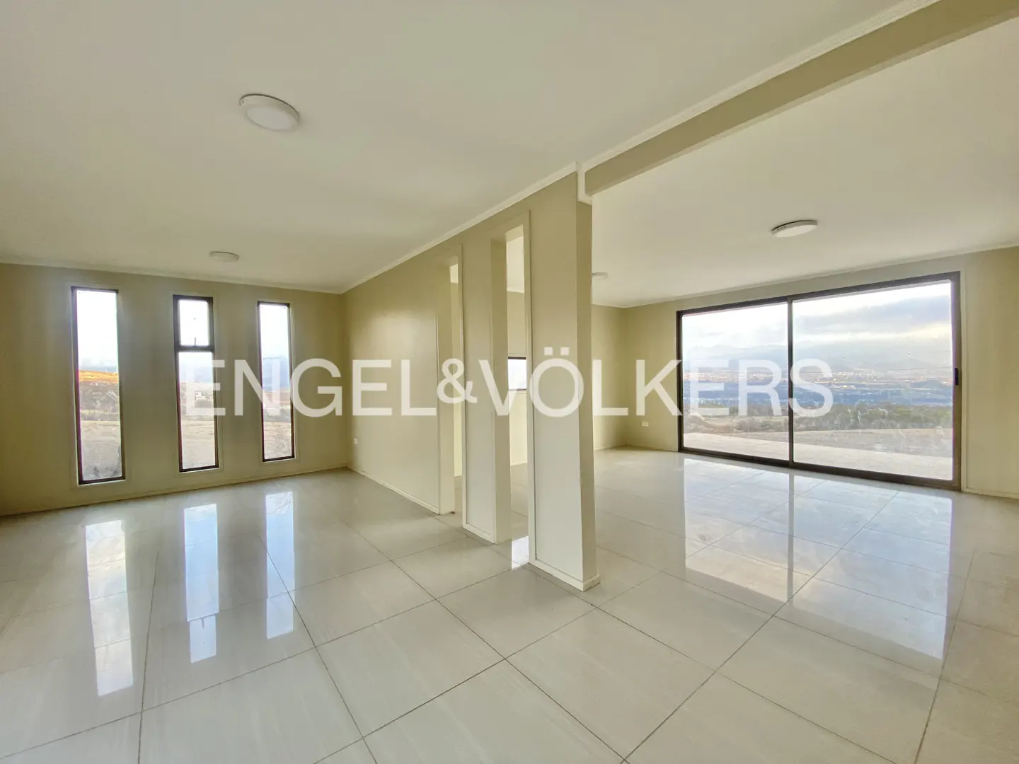 An empty, light-filled room with beige walls, white tile floors, and large windows with a view.