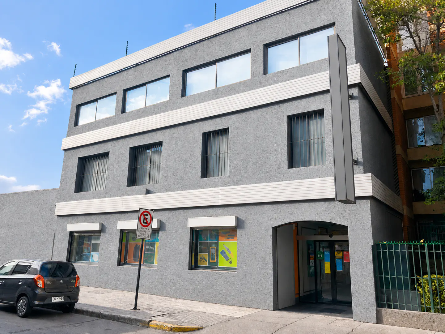 Three-story gray commercial building with a ground-level entrance, large windows, and a no-parking sign on the sidewalk.