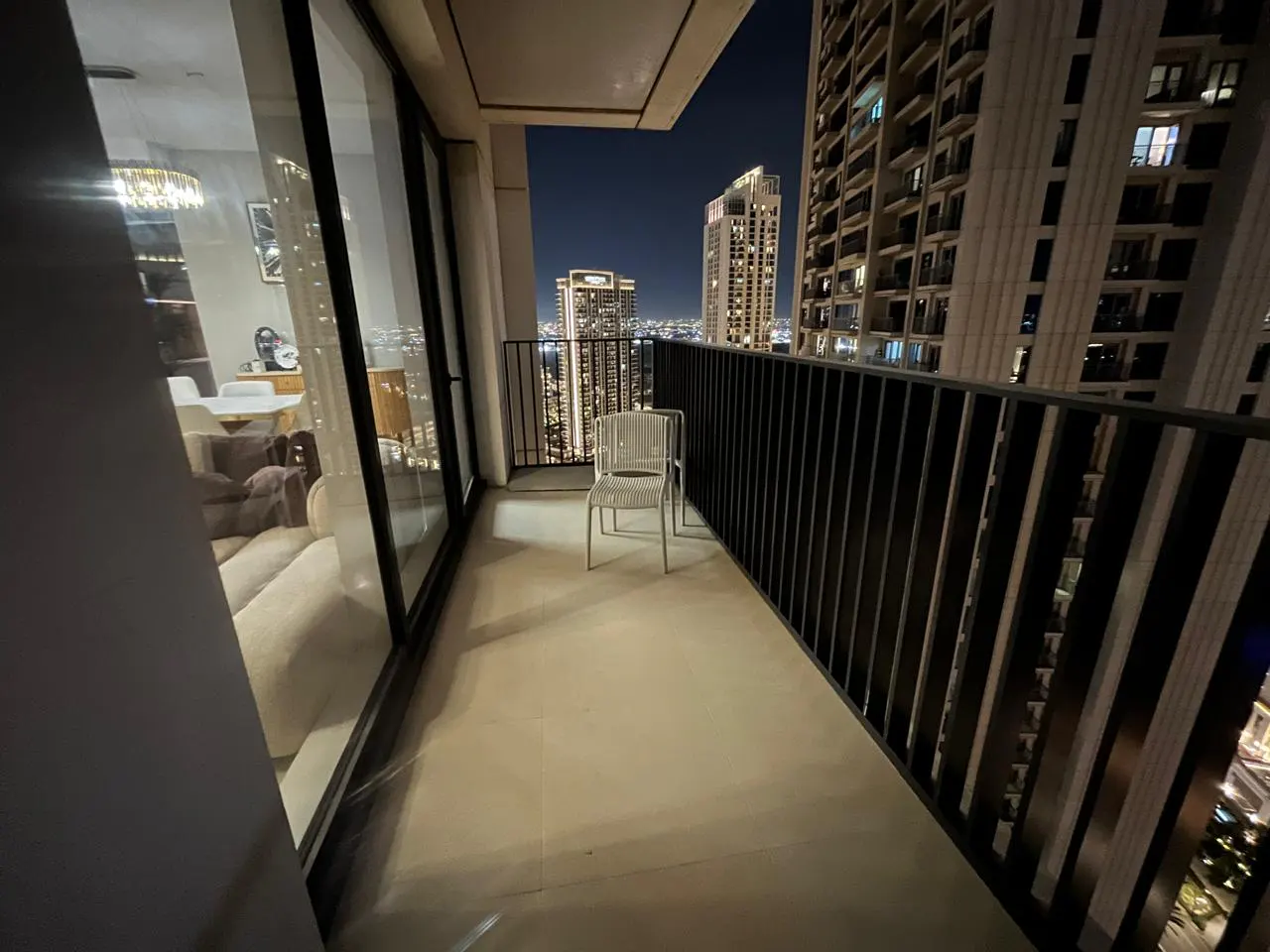 Balcony view at night with two white chairs, black railing, and city skyline in the background. Interior living space visible through glass doors.