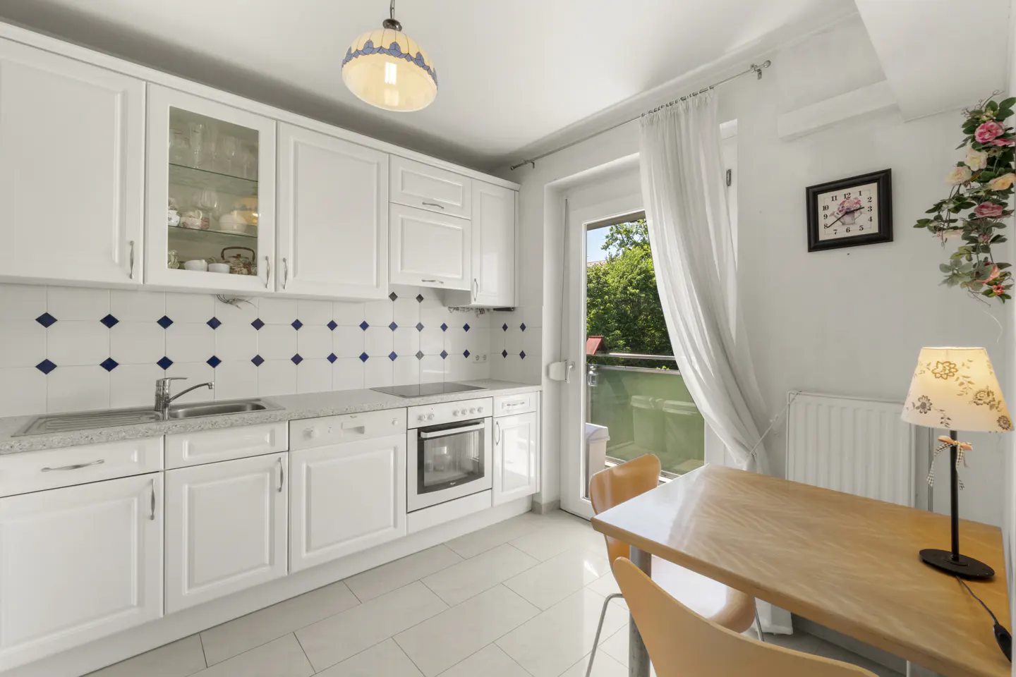 Bright kitchen with white cabinets, blue diamond backsplash, and light wood table with orange chairs near a balcony door.