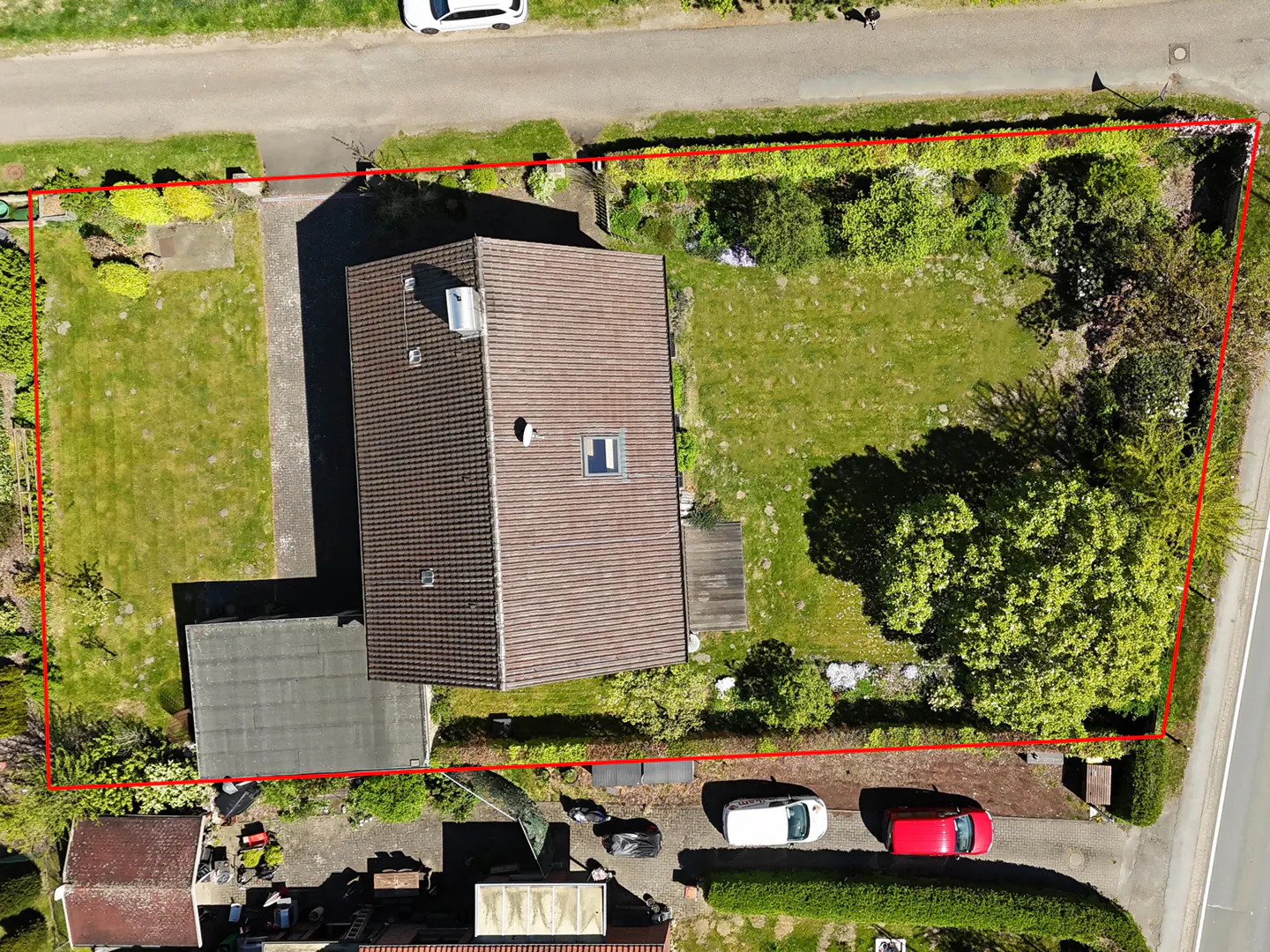 Aerial view of a house with a brown roof, surrounded by a green lawn and trees, outlined in red. Cars are parked nearby.