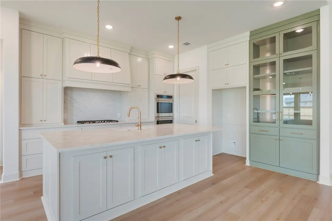 Bright kitchen with white cabinets, marble countertops, and a large island with a gold faucet. Green glass-front cabinets on the right.