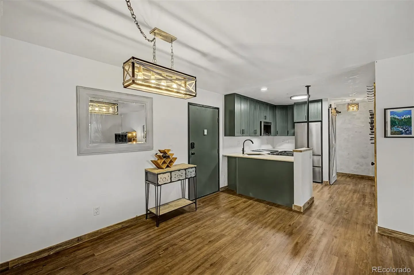 A bright, open kitchen and dining area with wood floors, green cabinets, and a modern light fixture. A decorative table sits near a mirror.