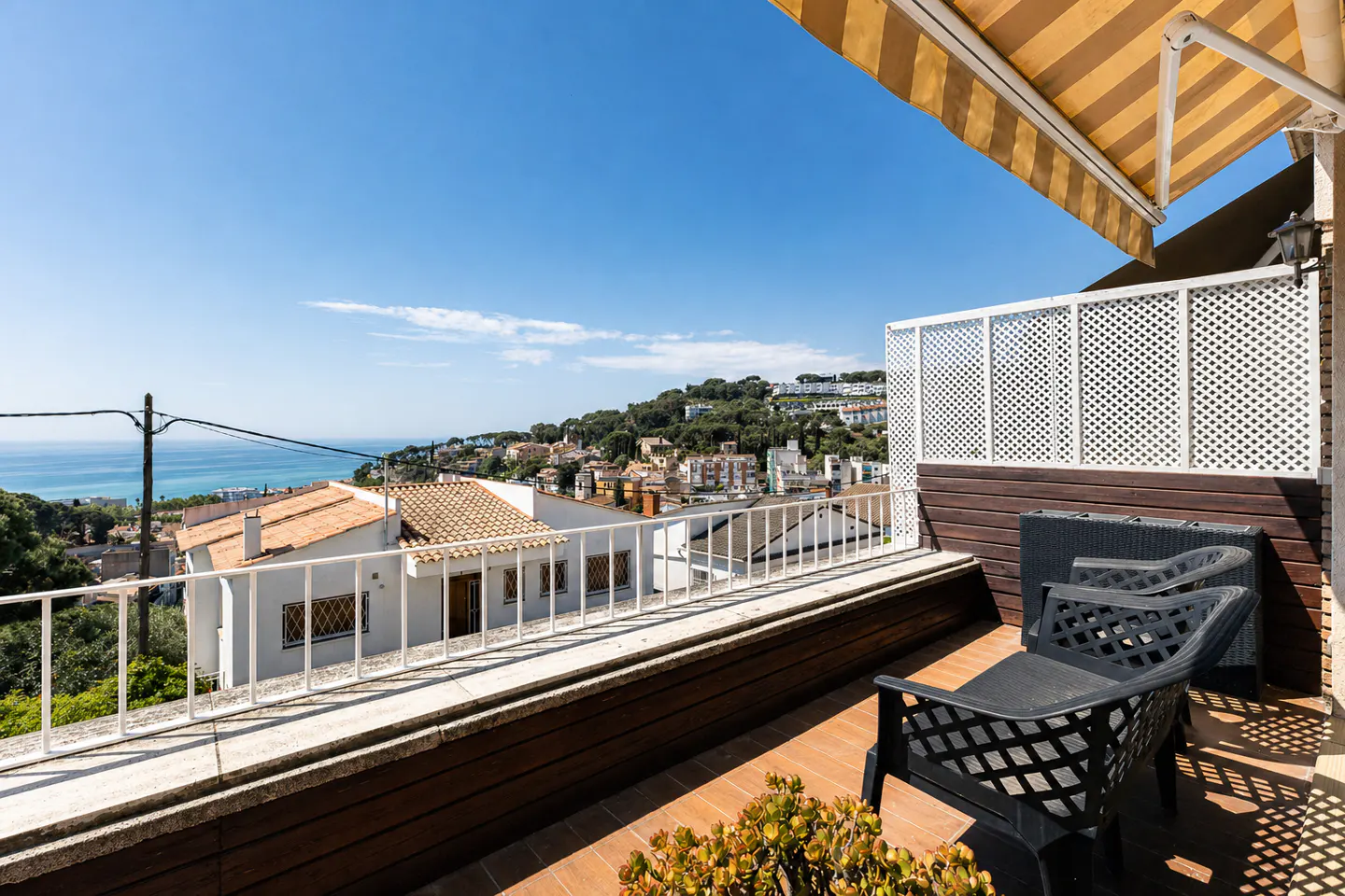 Balcony view with two black chairs, white lattice railing, and a striped awning. Distant buildings and blue sea visible.