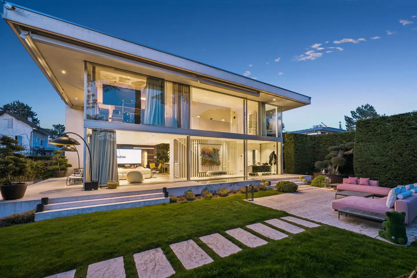 Modern two-story glass house at dusk with a landscaped lawn, stone path, and pink outdoor lounge furniture.