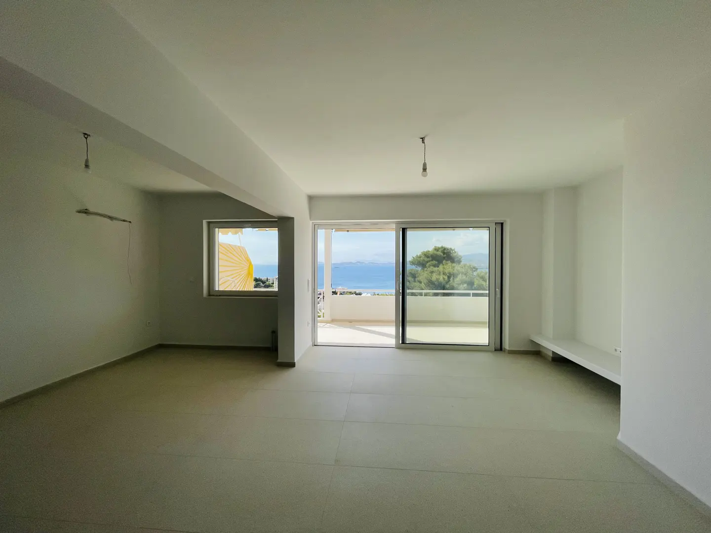 Bright, empty room with white walls, light gray tile floor, and ocean view through sliding glass doors to a balcony.
