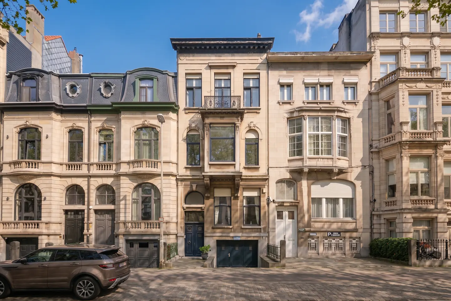 Row of beige stone townhouses with varying architectural styles under a blue sky. A brown SUV is parked in front.