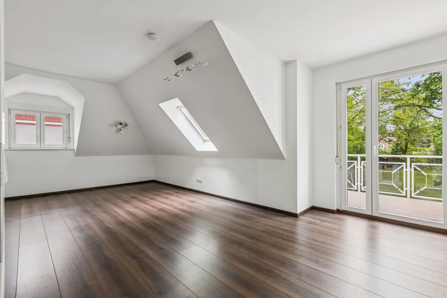 Bright attic room with dark wood floors, white walls, a skylight, and a window. Double doors lead to a balcony with a view of trees.