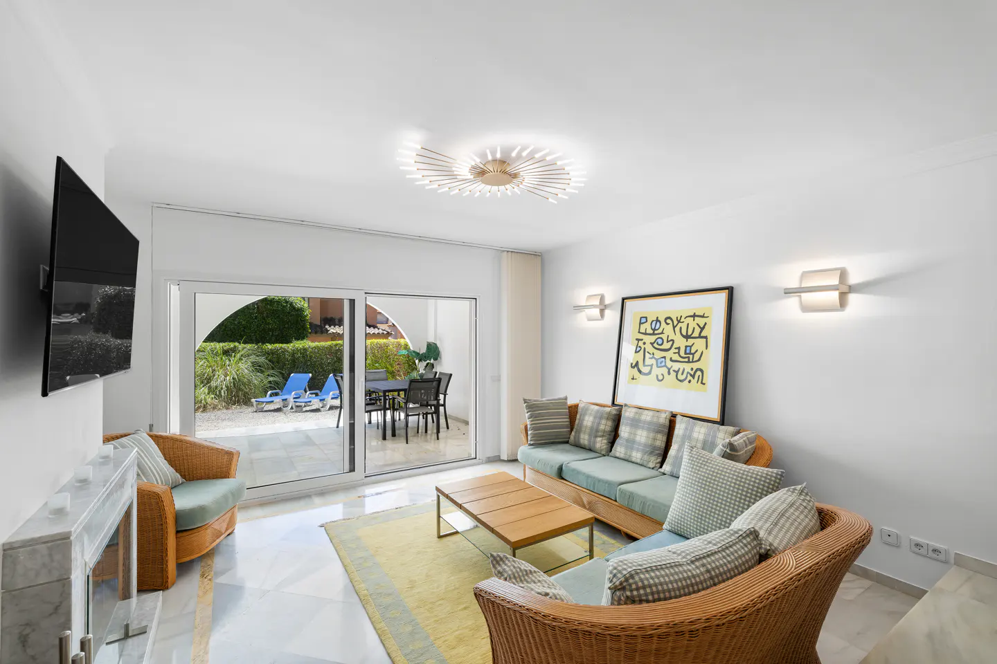 Bright living room with wicker furniture, a wood coffee table, and a yellow rug. Sliding glass doors lead to a patio with outdoor furniture.
