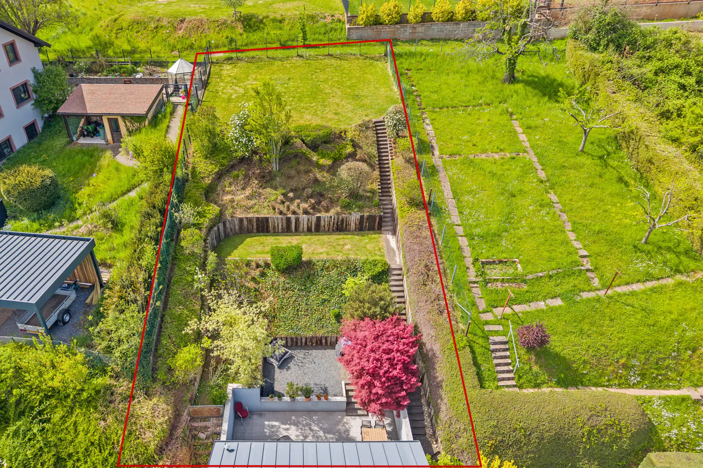 Aerial view of a property with a red border, featuring a green lawn, garden, patio, and a small building.