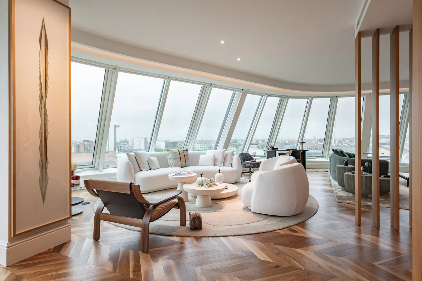 Bright, modern living room with curved windows overlooking a city. White sofa, chairs, and round tables on a circular rug. Herringbone wood floors.