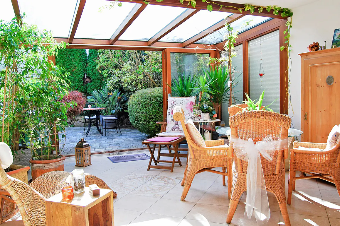 Sunroom with wicker furniture, glass roof, and open doors leading to a lush green garden patio.