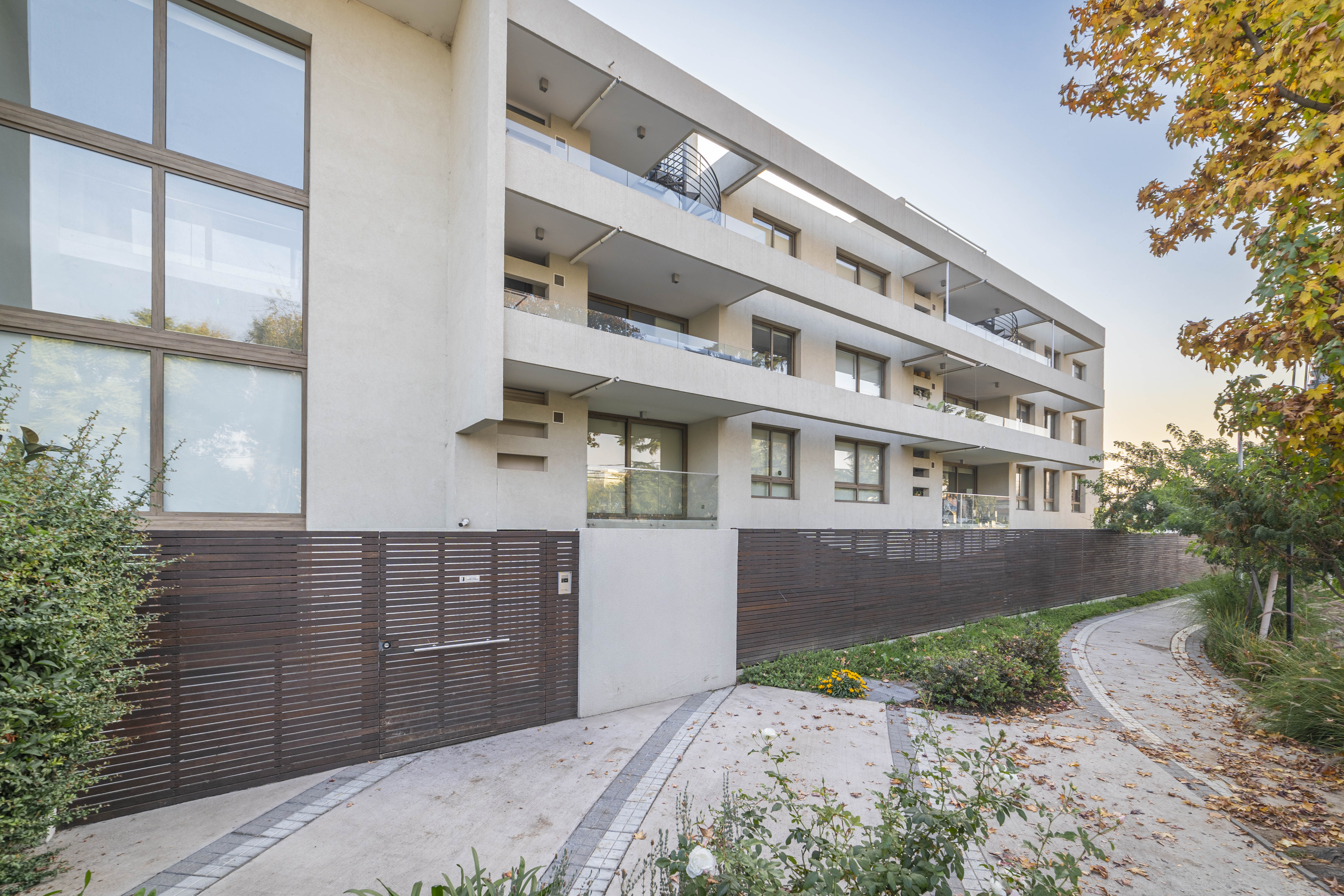 Modern apartment building with balconies and large windows, a brown wooden gate, and a stone pathway with trees.