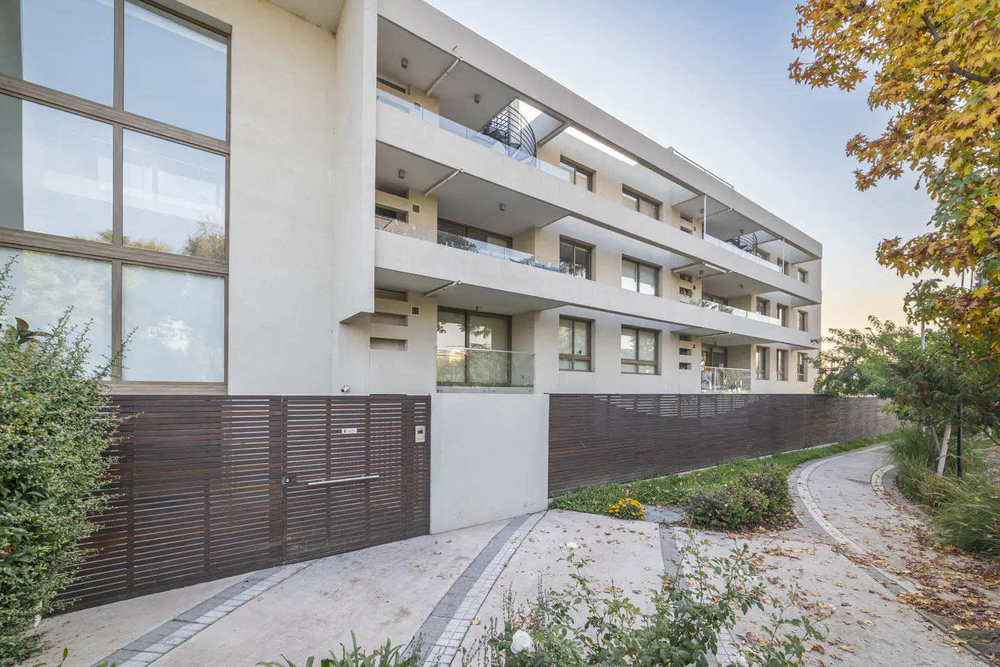 Modern apartment building with balconies and large windows, a brown wooden gate, and a stone pathway with trees.
