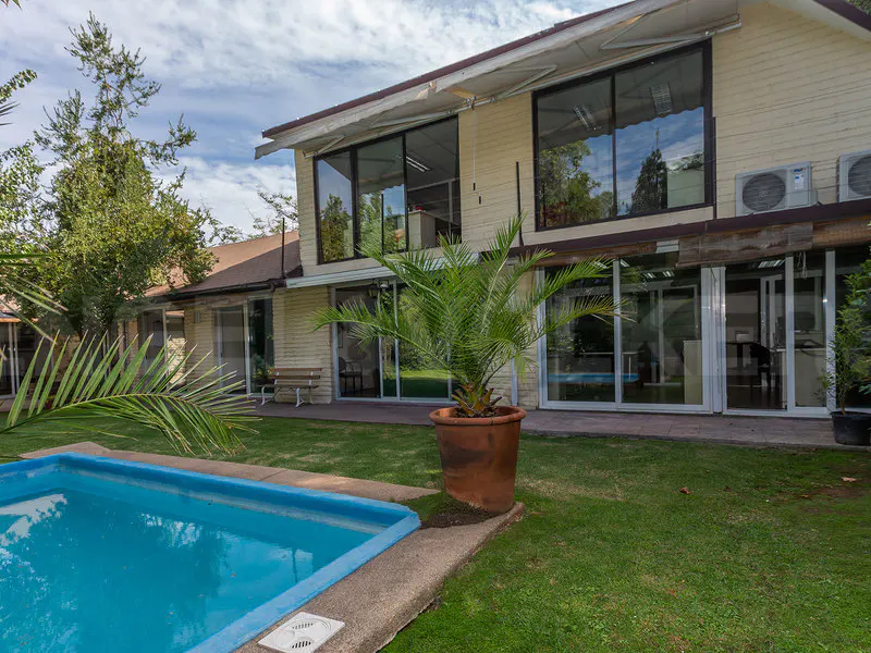 Backyard view of a two-story beige house with a blue swimming pool and a palm tree in a pot.