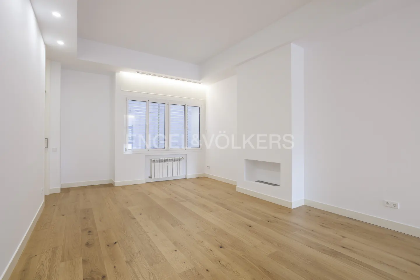 Bright, empty room with light wood floors, white walls, and a window with a radiator. A decorative fireplace niche is on the right wall.