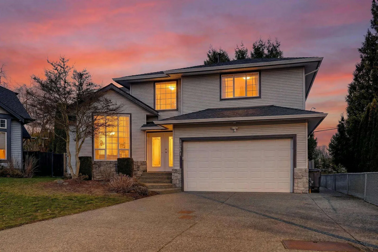 Two-story gray house with a white garage door and a pink and orange sunset sky.