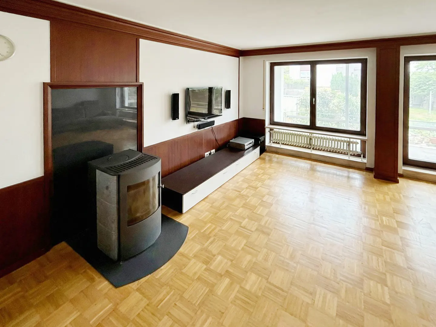 Living room with parquet floors, a wood-burning stove, a TV, and large windows. The walls are white with brown trim.