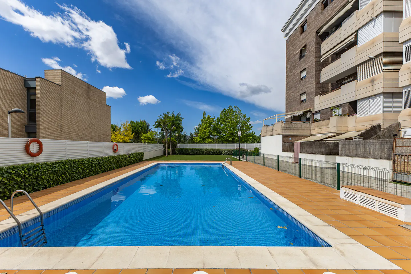 Outdoor pool with blue water and tile surround, next to a brick apartment building under a blue sky with white clouds.