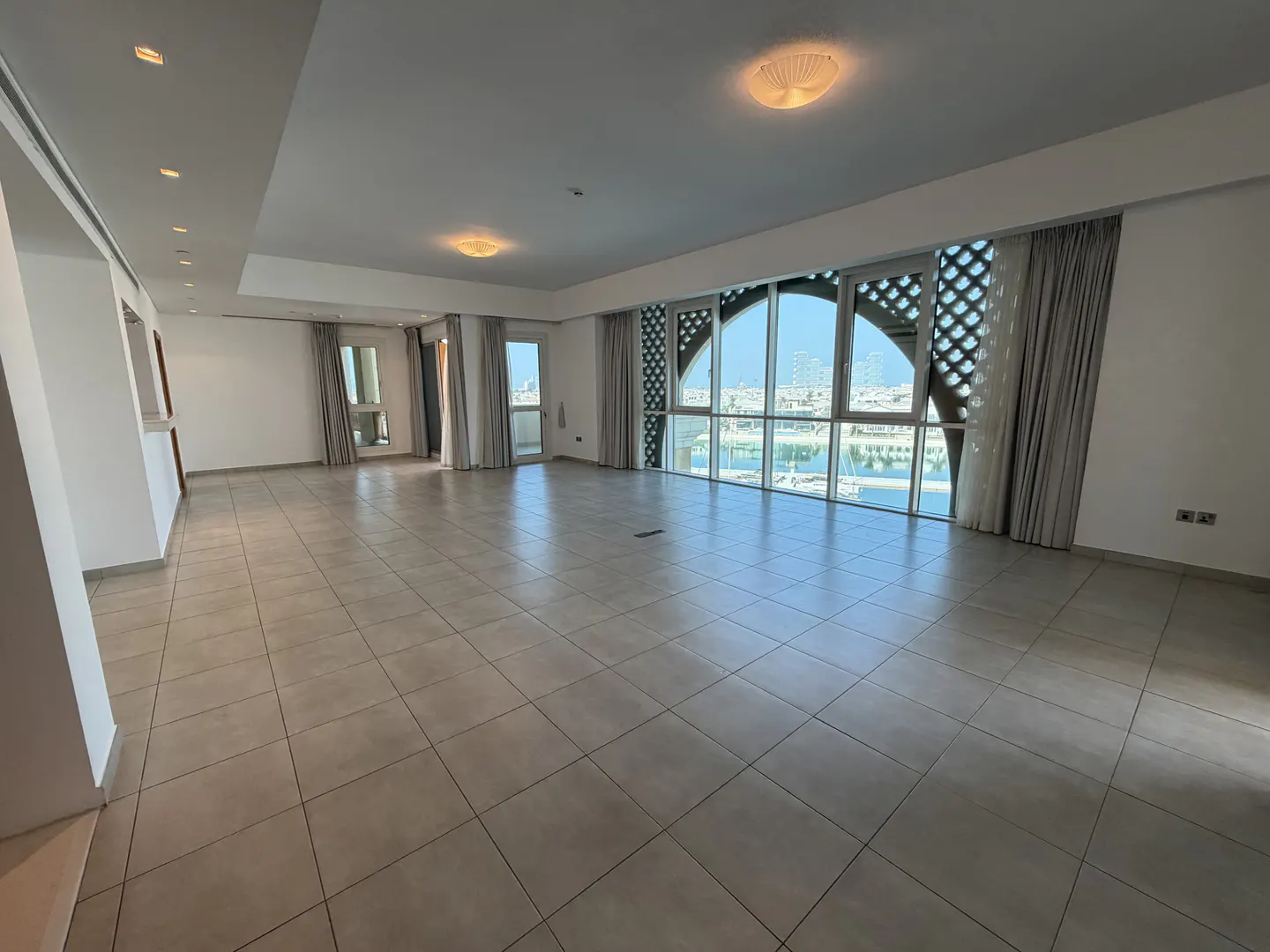 Spacious, unfurnished living room with light tile floors, white walls, and large windows overlooking a marina.