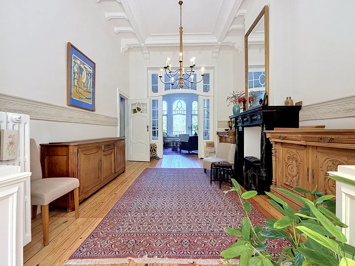 Grand foyer with wood floors, a red patterned rug, and white walls. A chandelier hangs from the ornate ceiling. A doorway leads to another room.