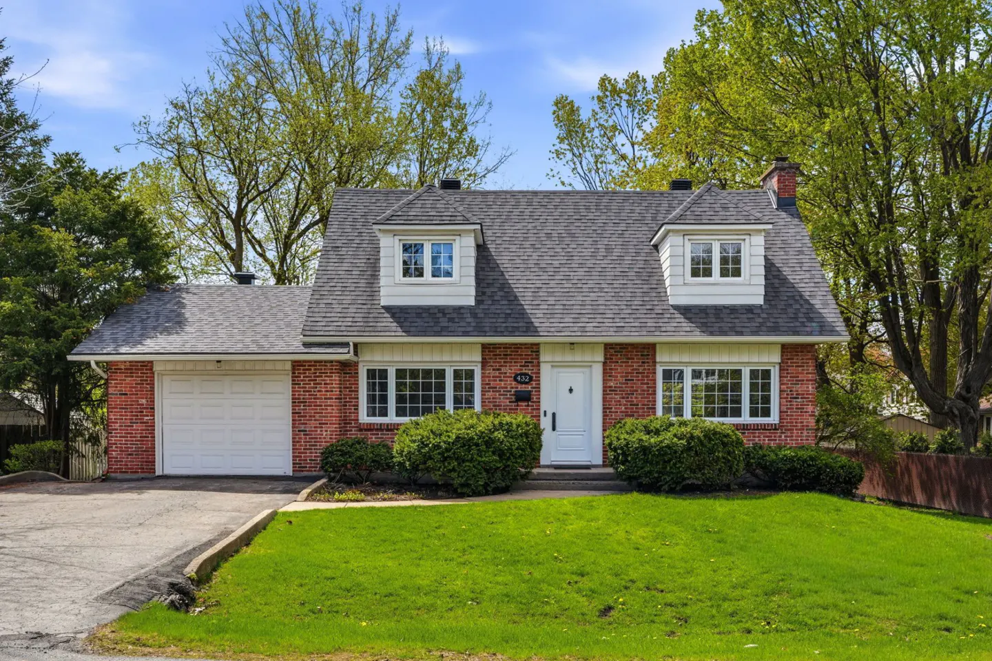 Red brick house with white trim, gray roof, and white garage door. Green lawn and trees surround the property.