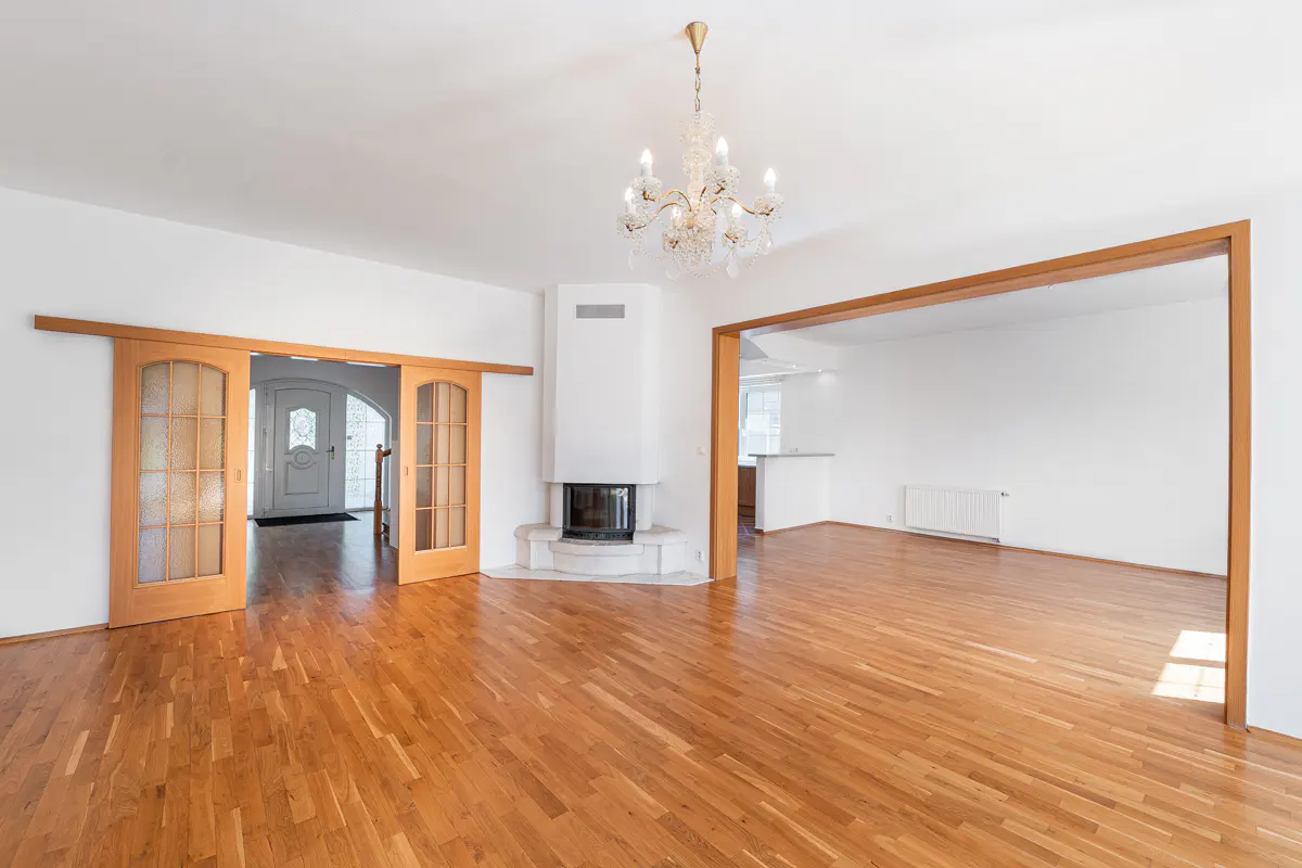 Bright, empty living room with hardwood floors, white walls, and a fireplace. A chandelier hangs from the ceiling. Sliding doors lead to the entrance.