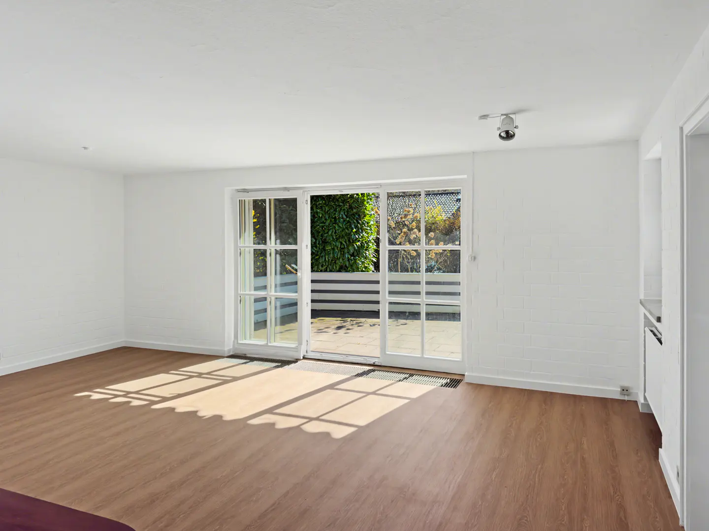 Bright, empty room with wood floors and white walls. Open glass doors lead to a patio with greenery. Sunlight streams in.